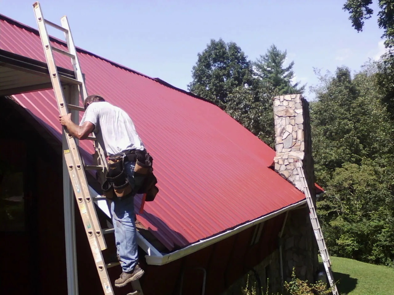 Roofer on a ladder beside a red metal roof and stone chimney — Soffit & Fascia Repair by Roofing Brothers in Ossipee